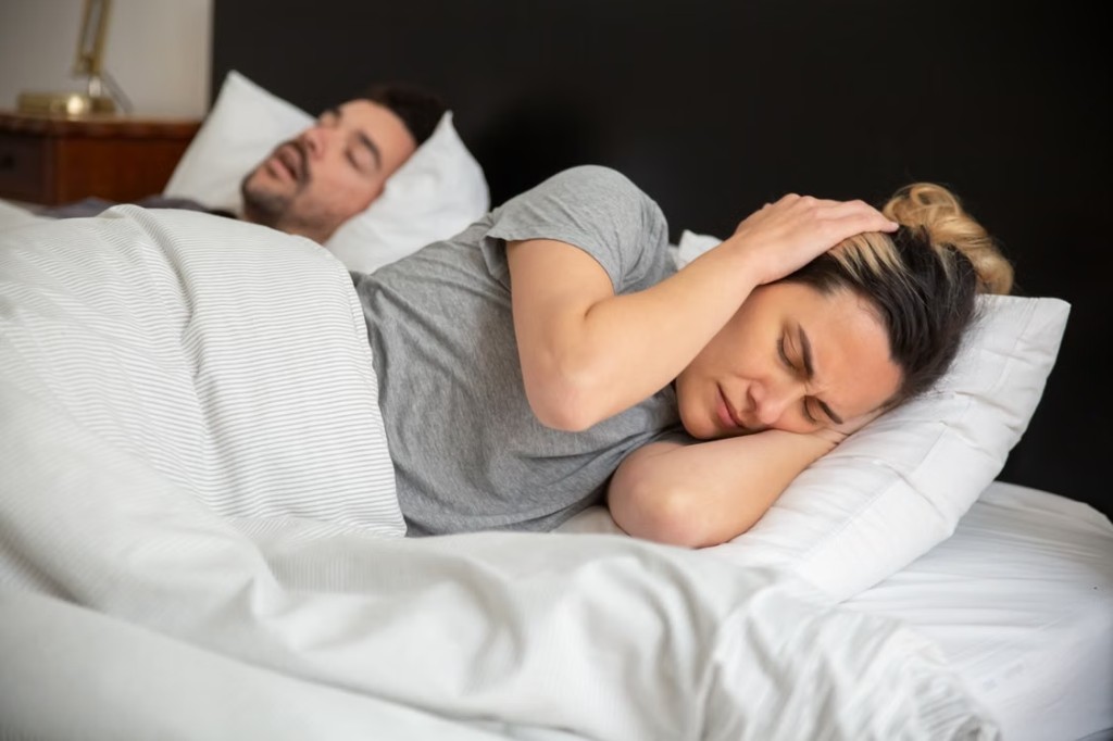 Woman covering her ear in bed while her partner snores beside her